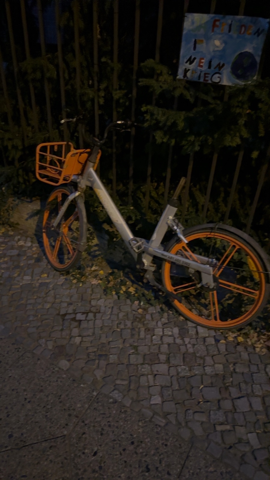 Ein beschädigtes Leih-Fahrrad mit orangenen Elementen steht an einem Zaun neben Gebüsch, daneben ein Schild mit Schrift, Christburger Straße 41, Pankow